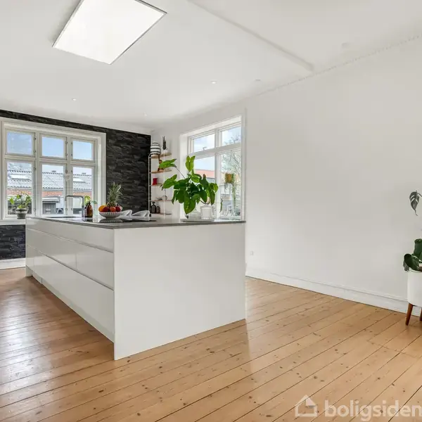 Køkkenø med planter og frugt på en træplankegulv i et lyst rum med store vinduer og en mørk kontrastvæg. En potteplante står på gulvet til højre.