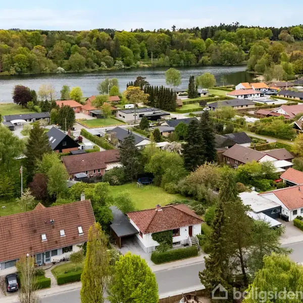 En dansk forstad viser huse med røde tage omgivet af grønne træer og haver. I baggrunden ses en sø med en skovet bakke. Træerne er frodige og grønne.