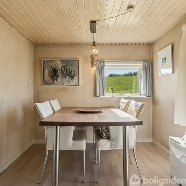 Et spisebord med fire stole står i et lyst rum med træloft. En lampe hænger over bordet. Vinduet i baggrunden viser en græsbevokset bakke udenfor.