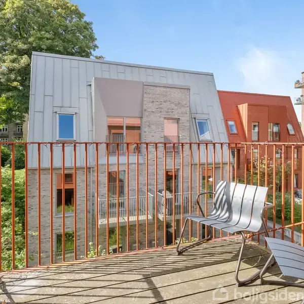 To moderne stole står på en balkon med et metalgelænder. I baggrunden ses moderne bygninger og frodige træer under en klar blå himmel.