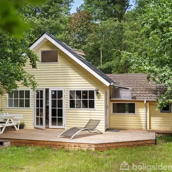 Et gult sommerhus med træveranda har liggestole og et havebord. Det ligger omgivet af frodig skov og grønne buske.