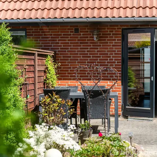 En have med et bord og to stole i flettet design står foran en murstensbygning med glasdør. Omgivet af blomster, grønne planter og et brunt hegn i solskinsvejr.