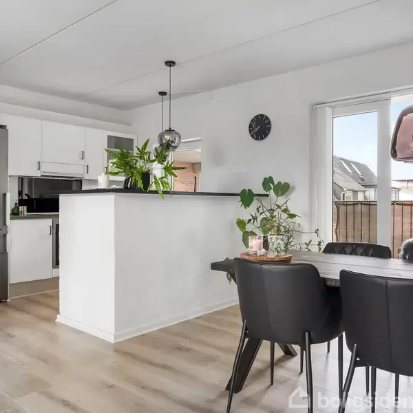 Køkken-alrum med spisebord fyldt med stearinlys og en plante. Køleskab og køkkenskabe til venstre. Store vinduer giver lys. Sort-rundt ur på væggen. Lyst trægulv.