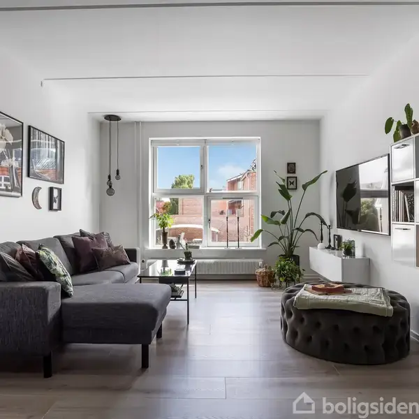 En stue med en grå sofa står ved siden af et lavt bord. Store vinduer bringer lys ind. Møbler som væghængte hylder og planter skaber en moderne atmosfære.