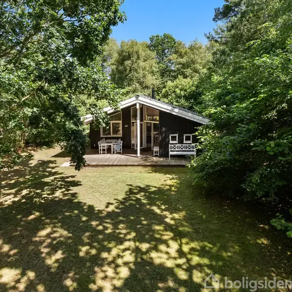 Et sort sommerhus, omkranset af træer, står stille på en græsplæne med en veranda foran, der har hvide havemøbler.