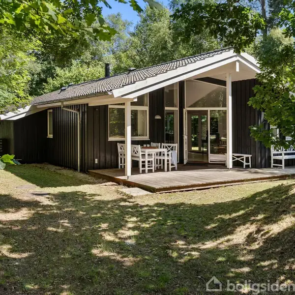 Sort træhus med skråt tag står stille omgivet af grønne træer. På terrassen foran huset er der et havesæt med bord og stole. Solrigt og skovomgivet miljø.