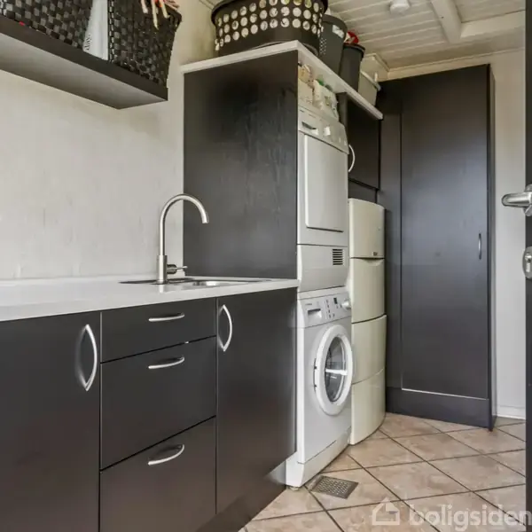 Vaskerum med vaskemaskine og tørretumbler stablet, omgivet af sorte skabe og opbevaringskurve på hvide vægge og lyse fliser. Dør til højre åbner ind i rummet.