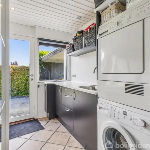 Vaskemaskine og tørretumbler står stablet i et moderne bryggers med skabe og vask. Rummet åbner ud til en terrasse via glasdør. Udsigt gennem dør til have.