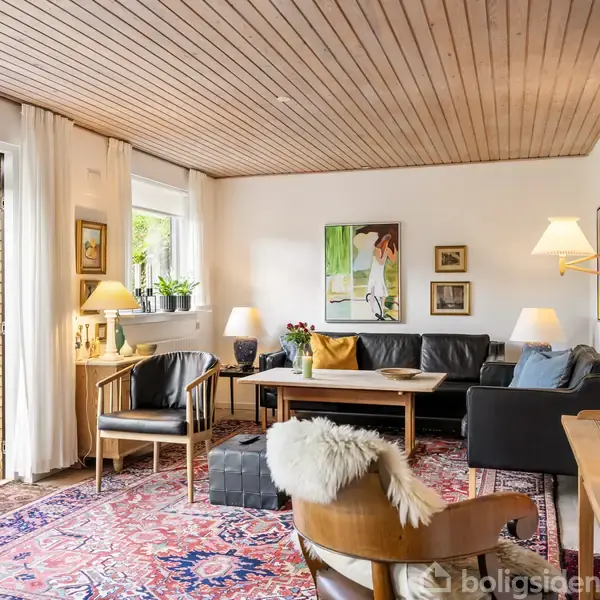 Stue med sofa og stole omkring et sofabord, dekoreret med farverige puder. Kunst på væggen, lamper tændt. Store vinduer, der åbner mod en terrasse i grønne omgivelser.