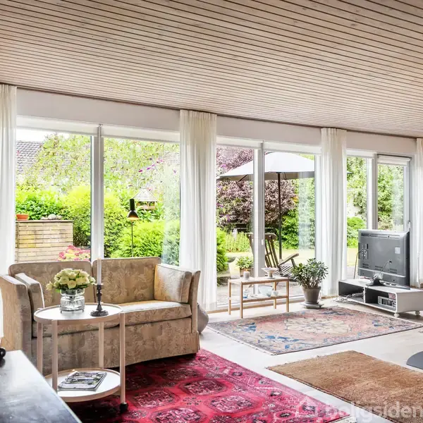 Sofaen står ved et rundt bord med en vase, foran store vinduer. Udendørs ses en have og terrasse med parasol og stole. Et fjernsyn står i hjørnet på gulvet.
