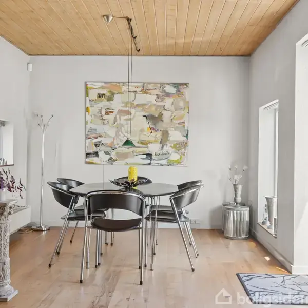 Et moderne spisebord med seks sorte stole står midt i et lyst rum. Væggen er dekoreret med abstrakt kunst, og store vinduer giver masser af naturligt lysindfald.