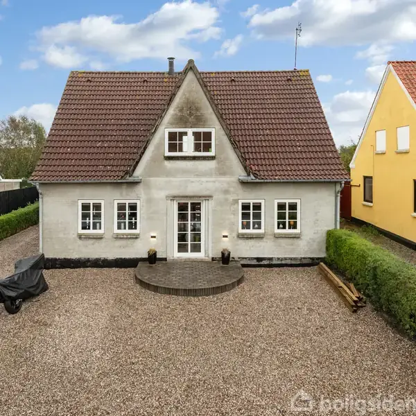 Et gråt hus med rødt tag står på en gruset forplads. Omkring huset er der hække og nabohuse i forskellige farver.