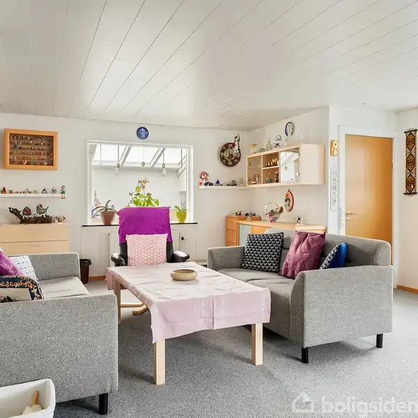 Stue med to grå sofaer omkring et bord med pink dug. Hylder med dekorationer og planter i vindueskarmen. Lyse vægge og loft. En dør fører til et andet rum.