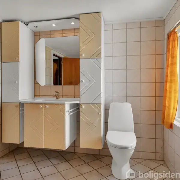 Et badeværelse med beige fliser, toilet og håndvask med indbyggede skabe. Et spejl hænger over vasken. Et vindue med orange gardiner giver naturligt lys.