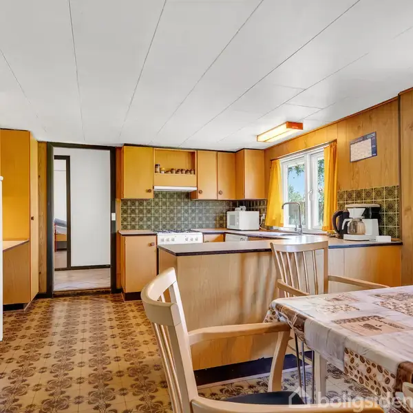 Køkken med træpaneler og mønstrede gulvfliser, indeholdende komfur, mikrobølgeovn og kaffeapparater. Et spisebord med dug og stole står ved vinduet med orange gardiner.