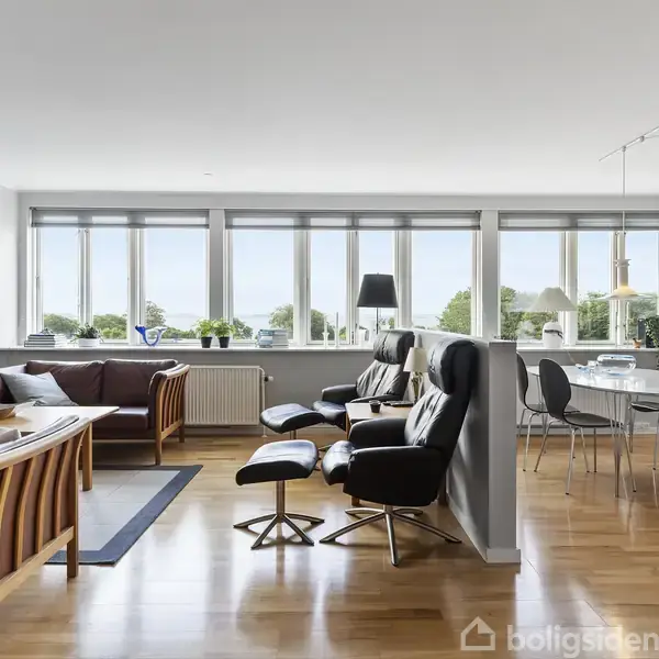 Stue med lædermøbler og spisebord stående på trægulv. Store vinduer langs væggen giver udsigt til grønt landskab udenfor. Elegant og lys indretning.