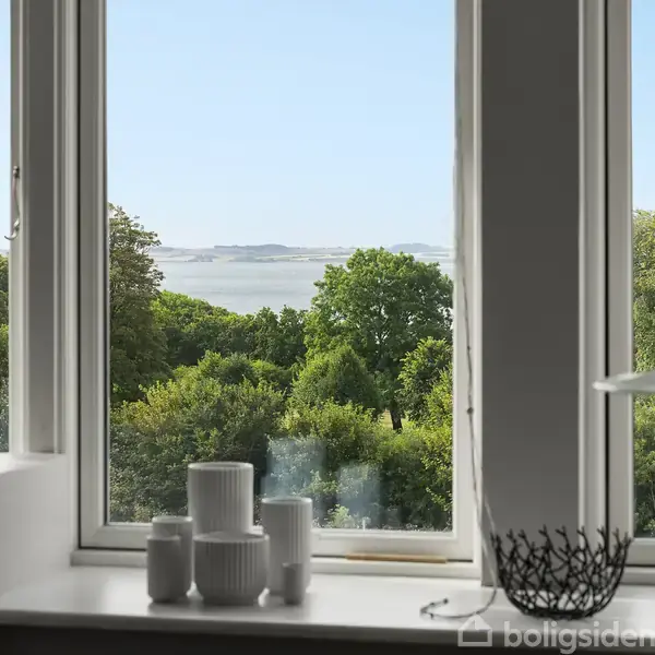 En vindueskarm med hvide porcelænsvaser og en elegant dekoration. Vindue med udsigt til frodig, grøn skov og en fjern horisont. Klar himmel og blå hav i baggrunden.