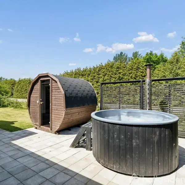 Trætønde-sauna står på en flisebelagt terrasse ved siden af et rundt, sort udendørs kar. Baggrunden består af grønne buske og en klar blå himmel.