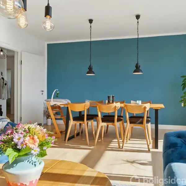 Et spisebord af træ med seks stole står i et rum med en blå væg. Pendellamper hænger over bordet. Rummet har planter, en blomstret vase og polstrede blå møbler.