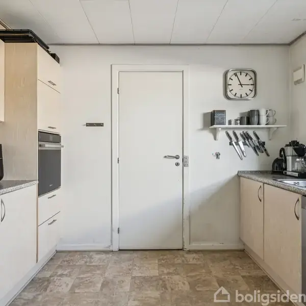 Køkken med hvide skabe og granitbordplader. Kaffemaskine og brødrister står til højre; ovn og airfryer til venstre. På væggen hænger et ur og knive.