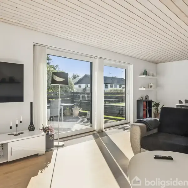 En moderne stue med en stor sofa, der står foran et fjernsyn. Store vinduer lukker naturligt lys ind, og udenfor ses en have. En spisestue er i baggrunden.