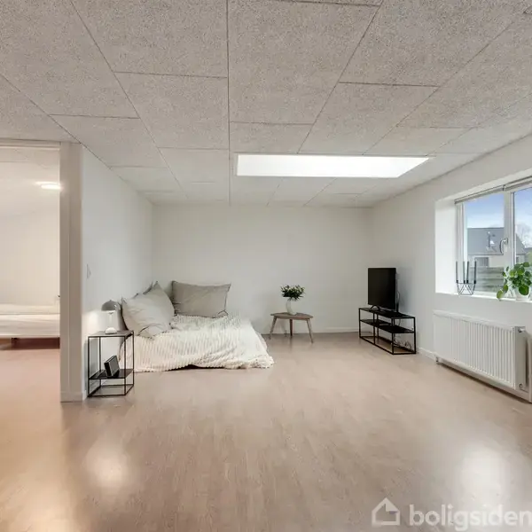 Et lyst og rummeligt værelse med en seng i venstre side, en lille sofa i midten, og et sort TV på et møbel til højre. Store vinduer lyser rummet op.