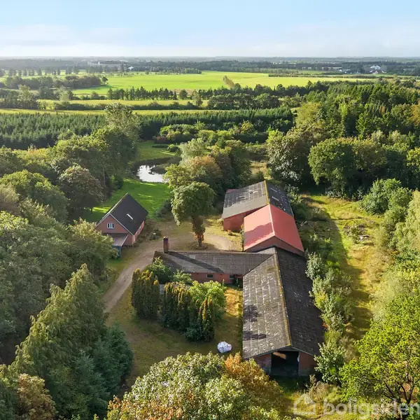 Gårdebygning med rødt tag ligger blandt frodige træer og grønne marker. En lille dam ses i nærheden, omgivet af skov. Landområdet strækker sig ind i horisonten.