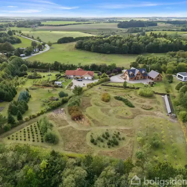 Luftfoto viser en stor ejendom med flere bygninger, omgivet af velplejede haver og grønne marker. Detaljerede haveanlæg ses med snoede stier og beplantning. Baghaven er skovklædt.