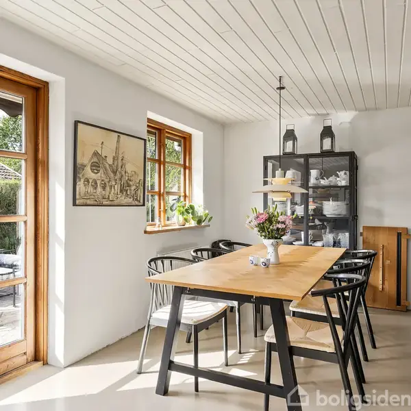 Et spisebord med blomsterdekoration i en lys spisestue. Stole er omkring bordet, og en vitrineskab står ved væggen. En dør åbner ud til en terrasse med havemøbler.