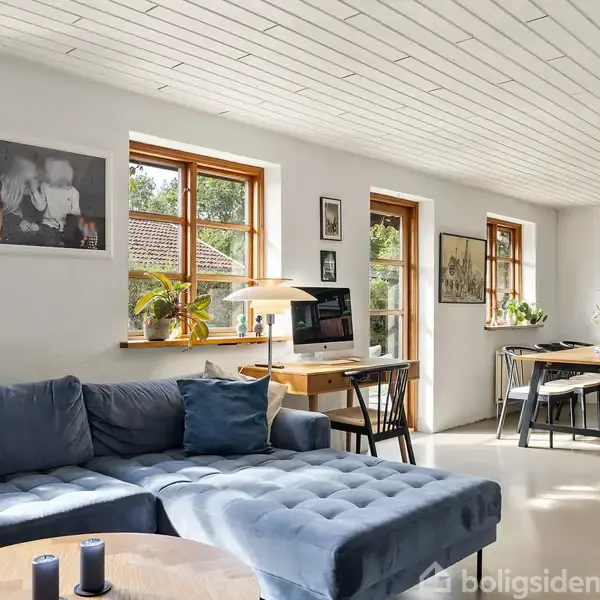 En blå sofa står i en lys stue med flere vinduer, der giver et skarpt lysindfald. Et spisebord er i baggrunden ved siden af en skænk, med dekorative planter og billeder på væggene.