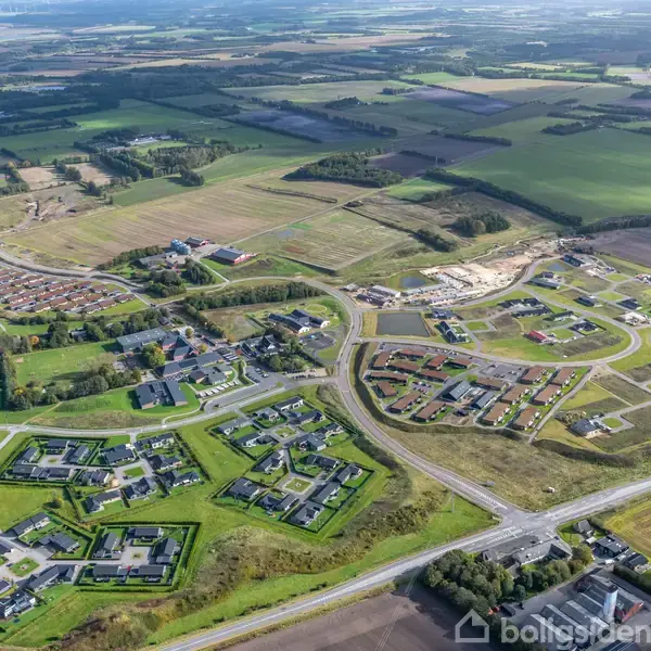 Boligområde med rækkehuse og parcelhuse, set fra luften omgivet af marker og skove. Veje forbinder beboelsesenhederne, mens grønne områder og små søer bryder landskabet.