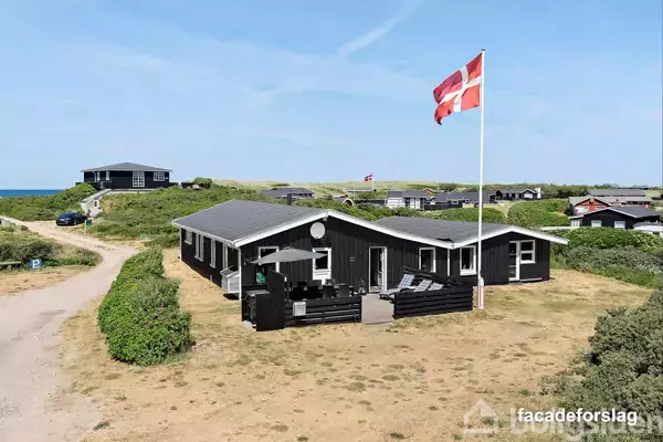 Et sort sommerhus står stille på en sandfyldt grund omgivet af lav pletvis vegetation. En dansk flagstang pryder haven, og kysten er synlig i baggrunden med flere huse omkring.