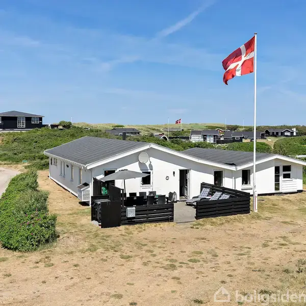 Et hvidt sommerhus står stille på en sandet grund med en stor, vajende dansk flagstang. Omgivet af andre huse og buske under en klar blå himmel.
