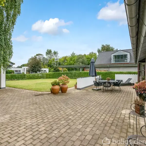 Stenbelagt terrasse med havebord og parasol i en stor baghave. Omgivet af grøn græsplæne og buske. Husfacade til højre og blå himmel med skyer i baggrunden.