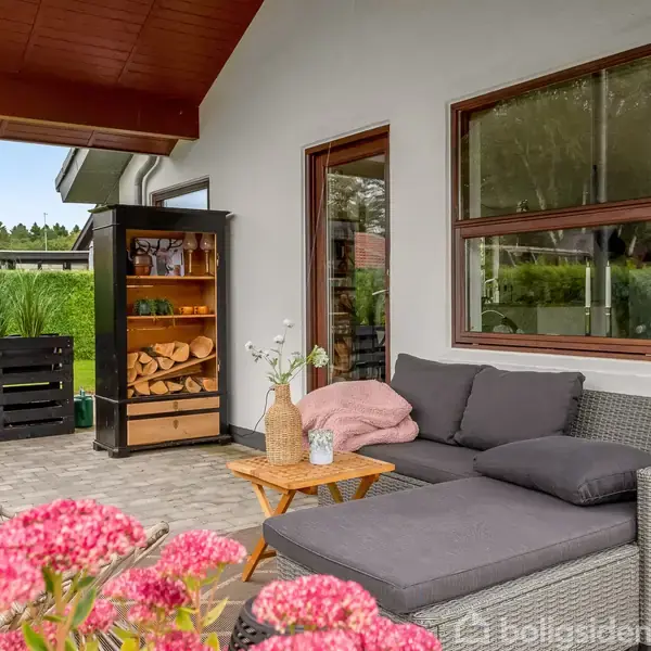 En grå udendørs sofa med lyserøde puder står på en terrasse. Ved siden af findes et lille bord med en vase. Bagved er en brændeovn og grønne omgivelser.