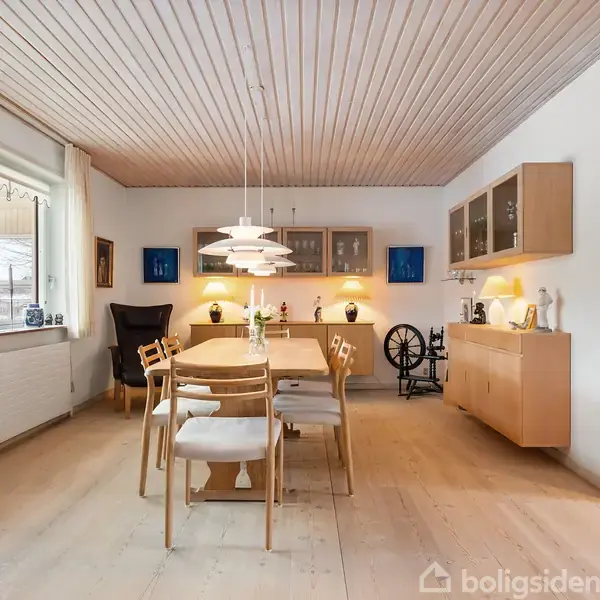 Spisebord med seks stole står i et lyst køkken-alrum. Over bordet hænger tre lamper. Til højre er skabe og en kamin, og til venstre store vinduer.