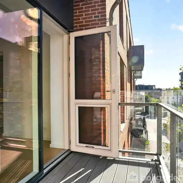Balkon med glasrækværk, åbnet glasdør ved en murstensbygning. Udsigt til andre bygninger og træer i sollys i en urban kontekst.