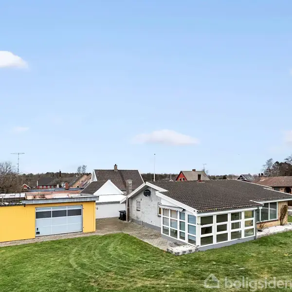 En enkelthus med garage og tilhørende gul bygning står på en stor græsplæne. Huset har et mørkt tag og mange vinduer, beliggende i et villakvarter under en blå himmel.