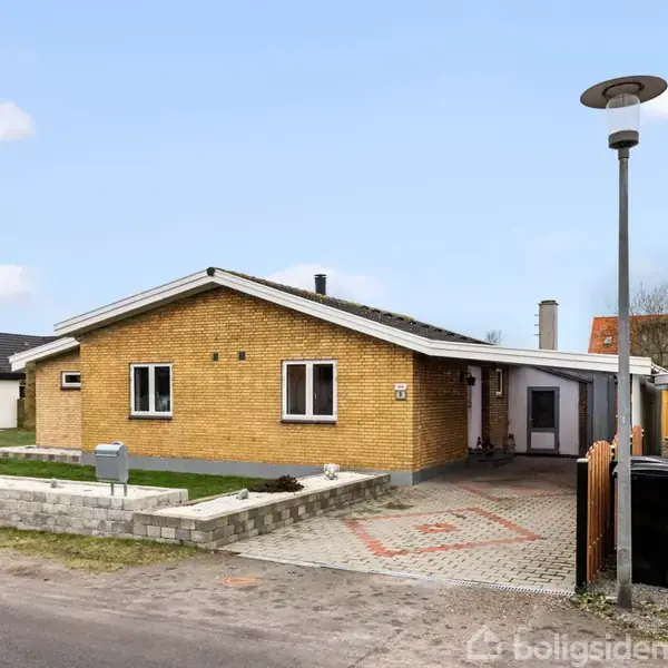 Et gulstenshus med fladt tag står på en flisebelagt indkørsel. En lille have med græsplæne og buske omgiver huset. Postkasse og stakit ses i forgrunden.