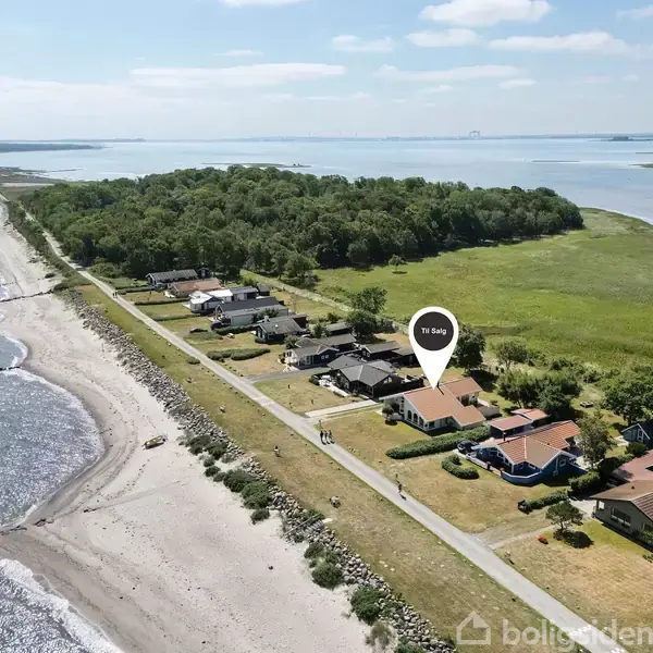 Et parcelhus ligger stille nær en kystlinje, markeret med en pin med teksten "15.495". Huset er omgivet af flere andre huse, grønne områder og en strand med udsigt over havet.