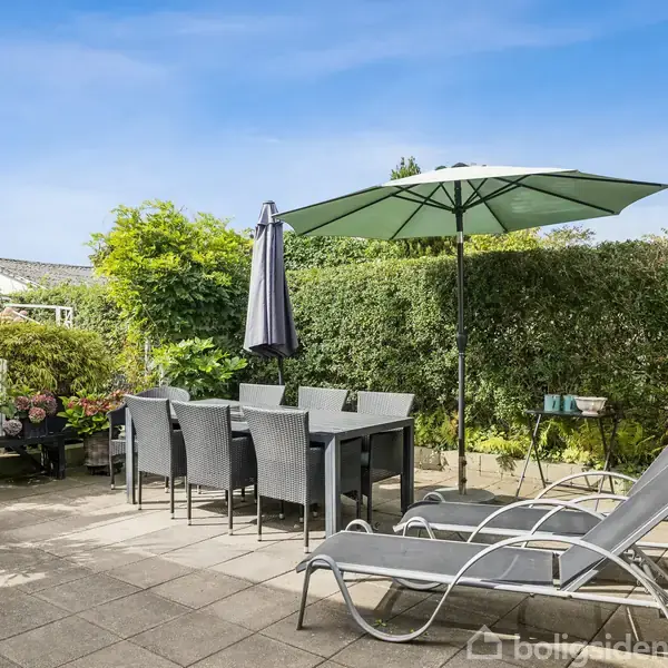 Terrassemøbler står på en flisebelagt terrasse. Et bord og stole er placeret under en stor grøn parasol. Liggestole er til højre. Haven er omgivet af frodig vegetation.