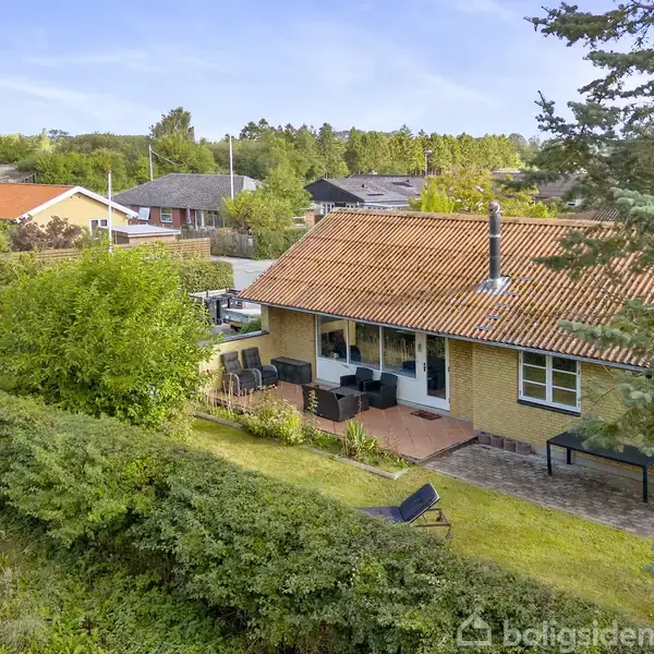 Et gulstenshus med rød tagsten, placeret i en grøn have med terrasse og havemøbler. Omgivet af buske og træer, med andre huse synlige i baggrunden.