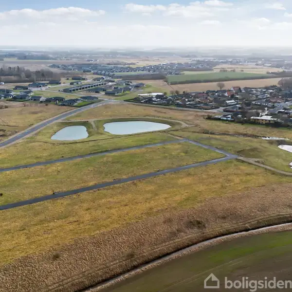 Grønt areal med tre små søer ligger ubenyttet, omkranset af stier og veje. Boligområde og marker strækker sig i baggrunden under en delvist skyet himmel.