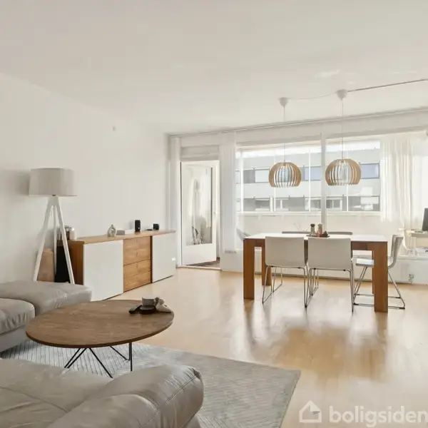 Stue med grå hjørnesofa og rundt sofabord til venstre, spisebord med fire stole og moderne pendellamper ved vinduerne til højre. Lyst trægulv og minimalistisk indretning.