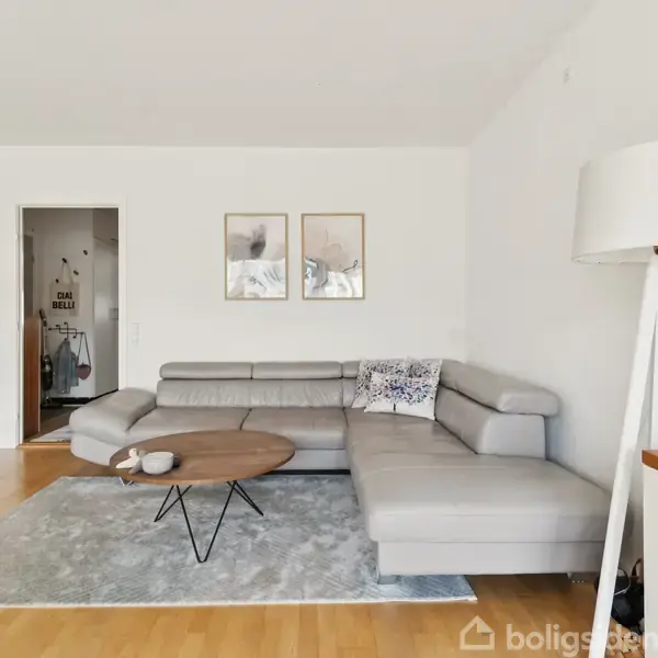 Grå hjørnesofa står på lys tæppe foran rundt sofabord i stue med hvidmalede vægge. To billeder hænger over sofaen, og en stor gulvlampe står til højre.