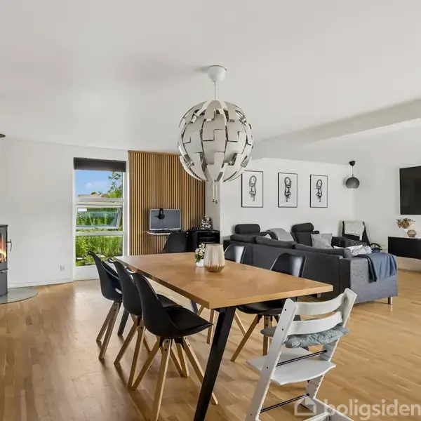 En stue med spisebord har seks stole omkring det og en dekorativ lampe over. Til venstre står en brændeovn tændt, og til højre er der sofaer, et stort TV, og en vægdekoration.