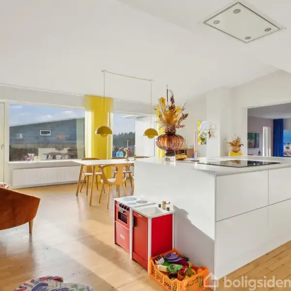 Et moderne køkken med køkkenø er centralt placeret, omringet af en spisestue med spisebord og stole. Der er store vinduer med gule gardiner i et lyst rum.