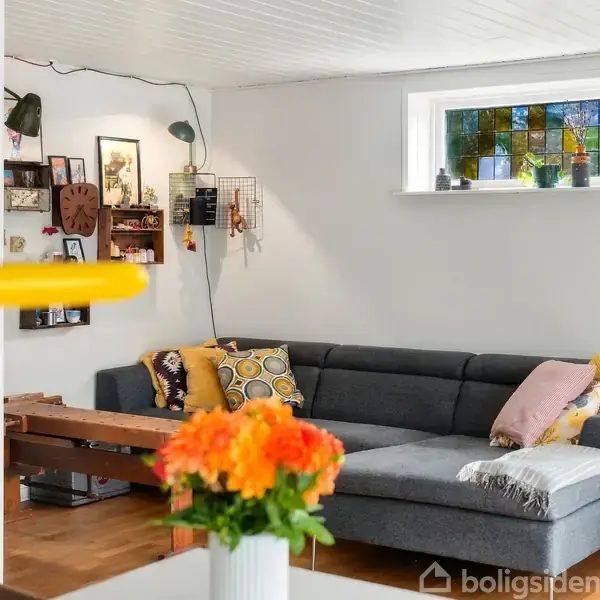 Grå hjørnesofa med farverige puder står i stue med hvide vægge. En blomstervase med orange og gule blomster står på bordet. Et lille vindue har farvet glas.