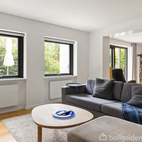 Stue med grå sofa og sofabord, beliggende i et lyst rum med to store vinduer og grøn udsigt. I baggrunden ses et spiseområde med stole og en bogreol.