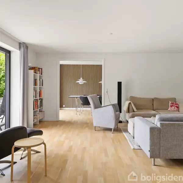 Stue med trægulv indeholder en grå sofa, sort lænestol og rundt bord. I baggrunden ses en spiseplads med bord og stole. Store vinduer åbner ud mod en terrasse.
